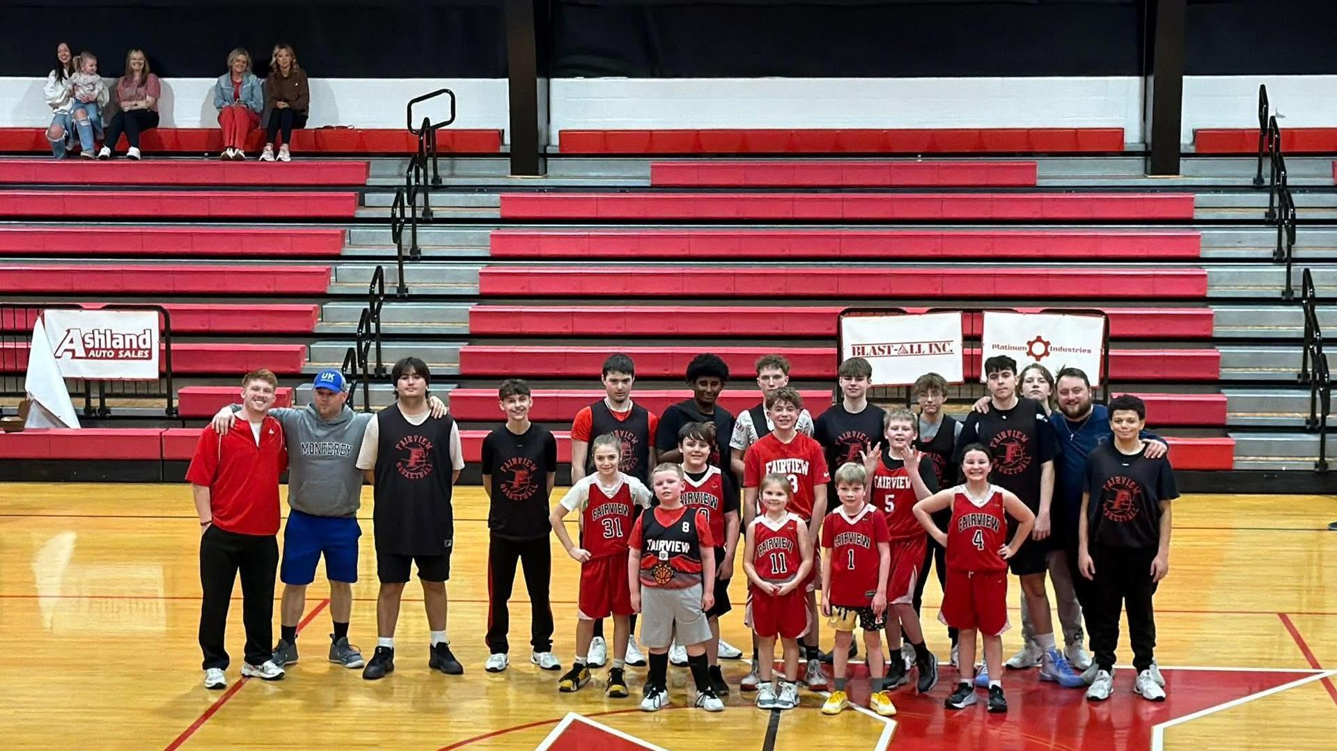 A group of basketball players posing together on a gym court with spectators in the background.