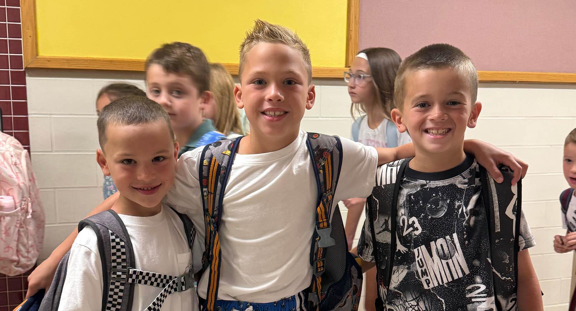 Three boys smile together in a school hallway, with other students visible in the background.