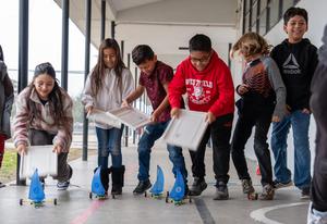 Elementary students line up outside, racing their LEGO-built sail cars by using plastic bin lids to create wind. The students are smiling and fully engaged, showing teamwork and excitement during the STEAM activity.