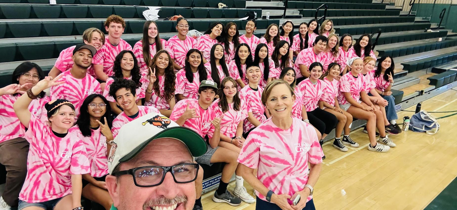 A large group of students in pink tie-dye shirts poses for a group photo on bleachers.
