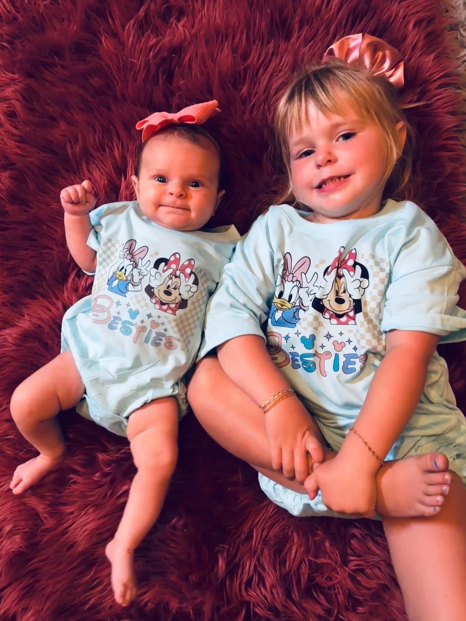 Two young girls in matching outfits smiling on a fluffy rug.