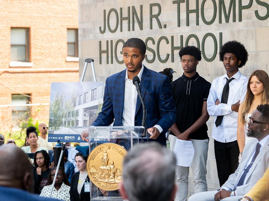 A young man speaks at a podium outside John R. Thompson High School, with an audience behind him.