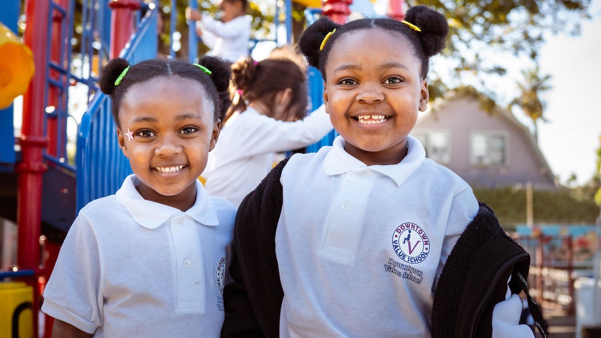 Two smiling girls with pigtails standing in front of a playground.