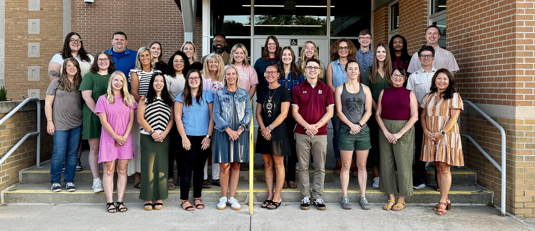 three rows of staff stand smiling on the front steps of East Elementary School with their hands clasped in front of them