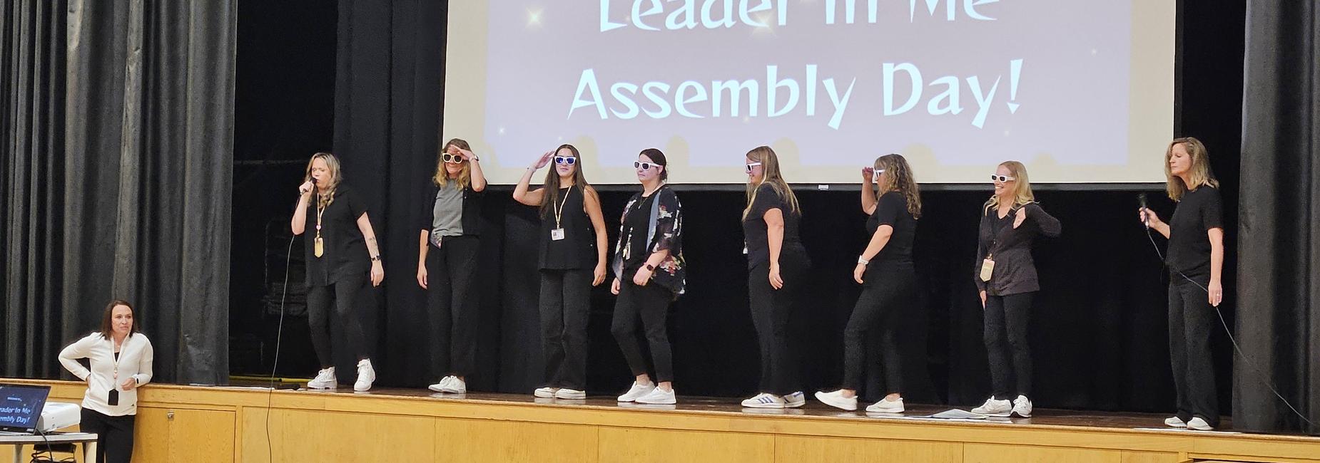 A group of people performing on stage at an assembly, wearing sunglasses and casually dressed.  Leader in Me Morning Assembly ~ Logan is looking for Hallway All-Stars!