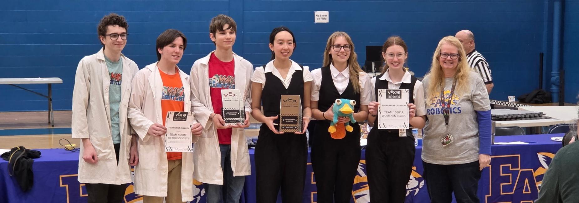 A group of eight students celebrates with trophies and certificates at a robotics competition.
