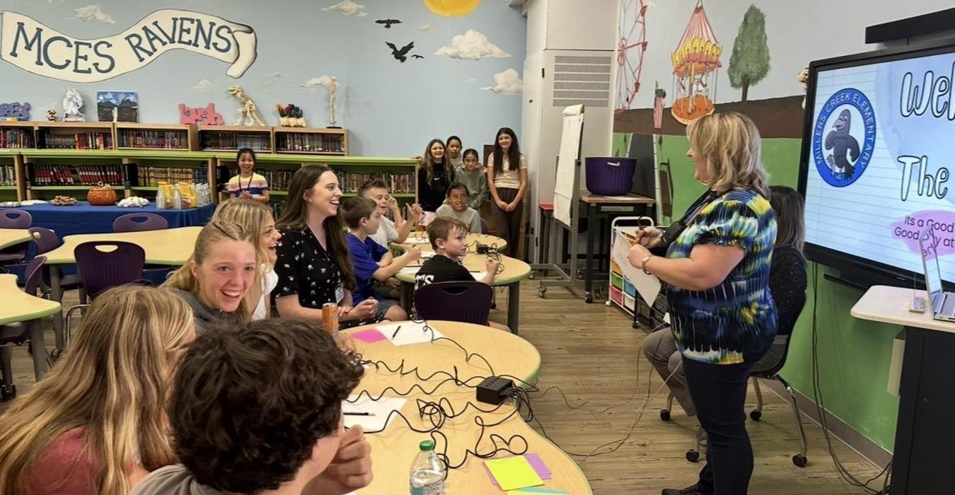 A group of students and adults engaged in a classroom discussion with a presentation screen.