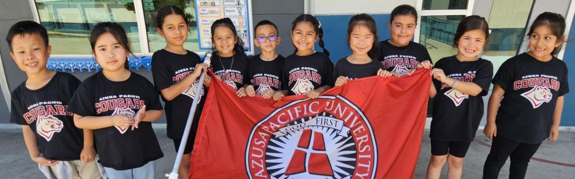 Group of children holding an Azusa Pacific University flag outdoors.