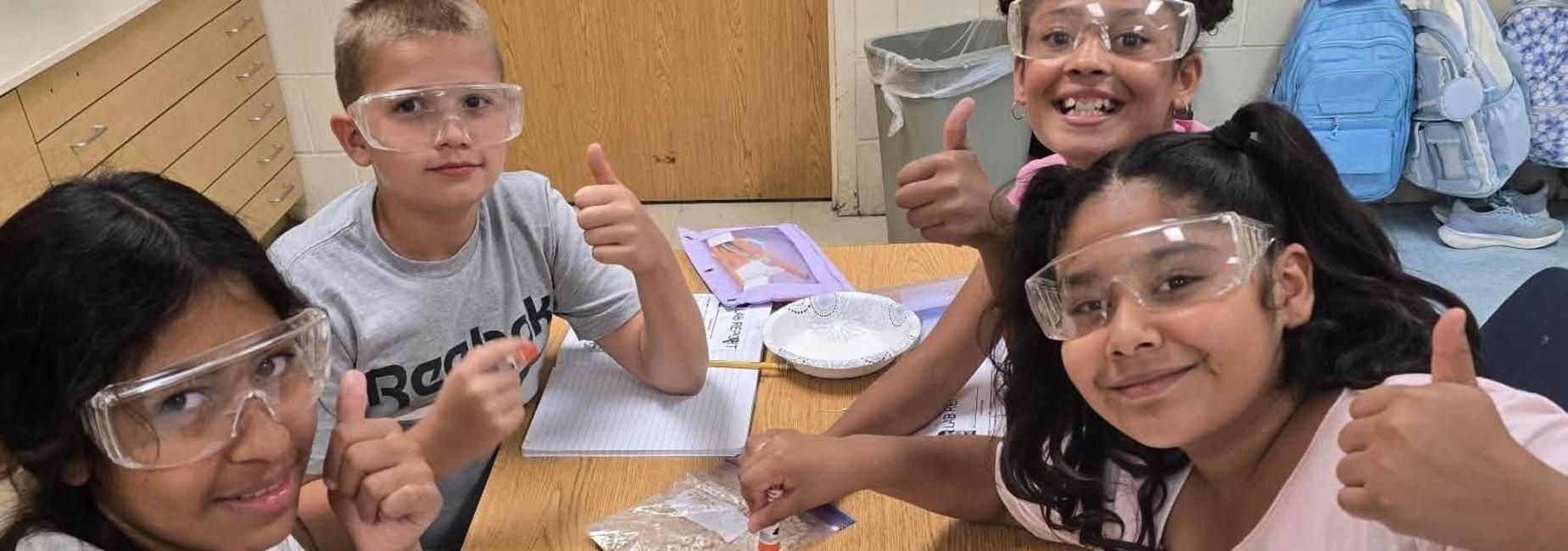 Group of children in safety goggles giving thumbs up while exploring a science experiment.