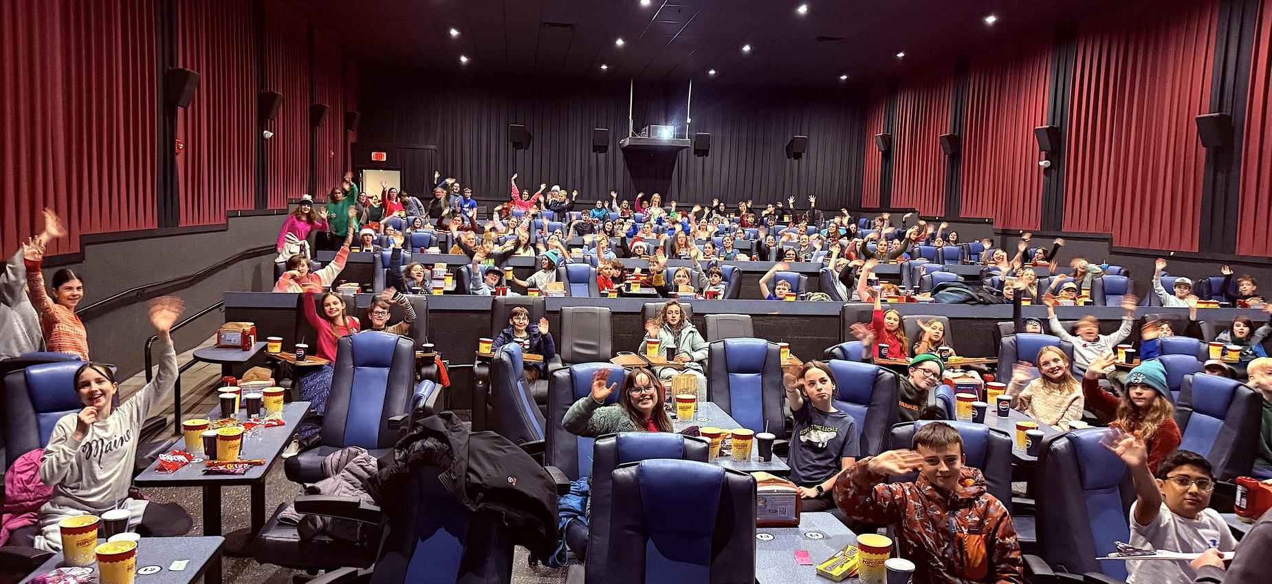 A large group of children and adults waving from their seats in a movie theater.