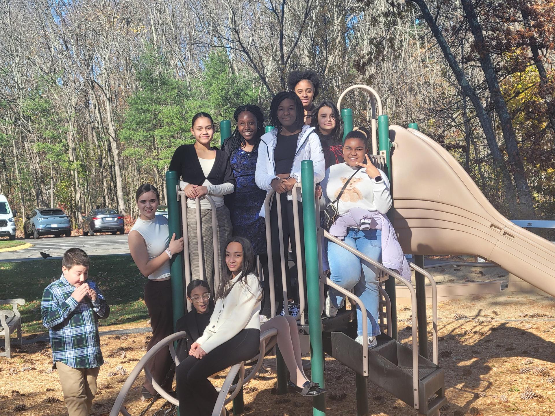 A group of diverse children and teenagers are gathered on and around a playground slide structure on a sunny day. They are smiling and interacting, suggesting a cheerful team or club outing.