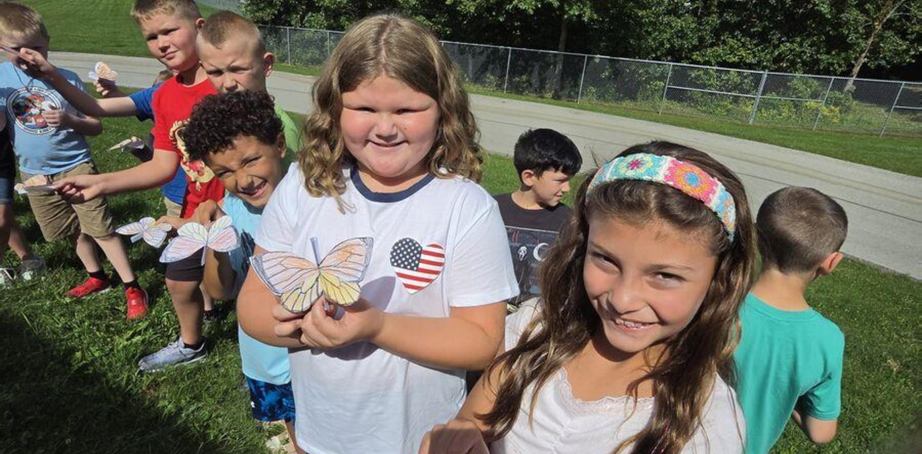 Children holding handcrafted butterflies outside in a sunny setting.