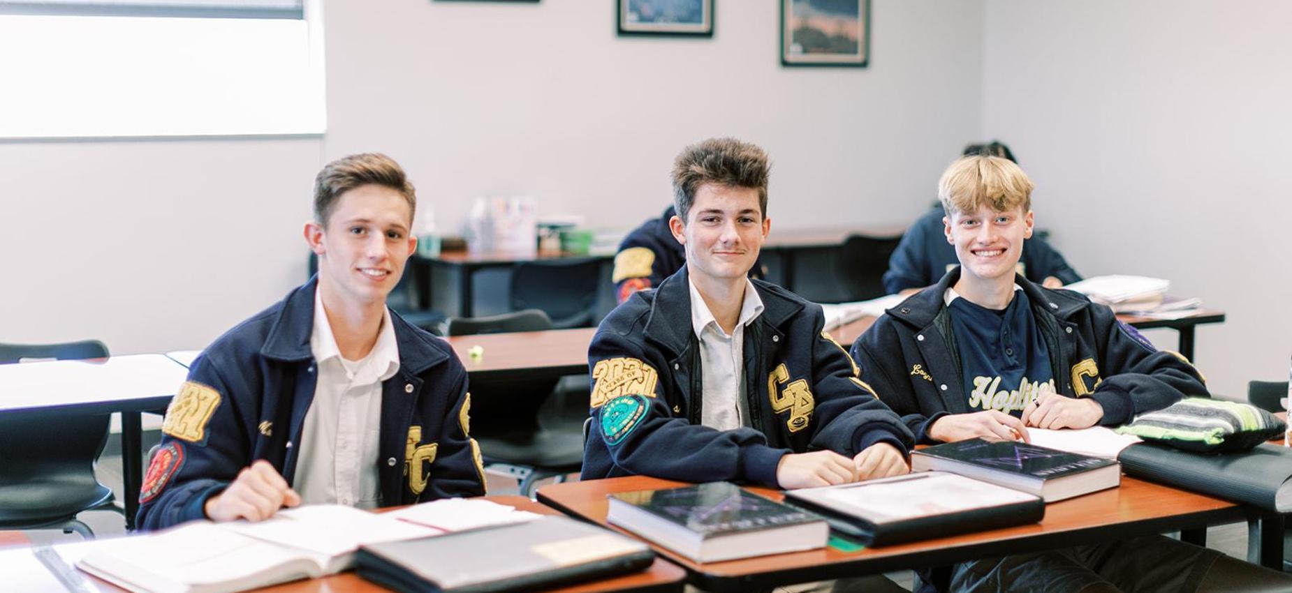 Three smiling high school students in navy blazers sit at their desks