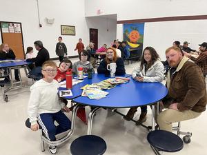 Group of children and adults sitting at a blue table in a school cafeteria.