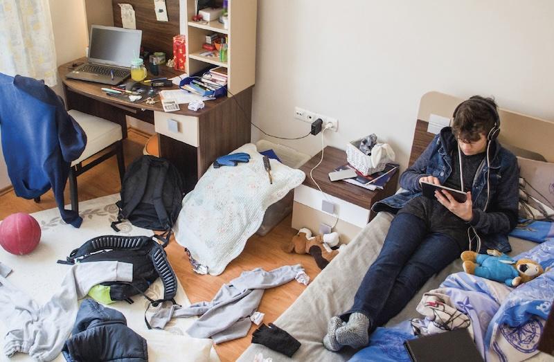 Older student listening to music and on an iPad in a disorganized, messy room