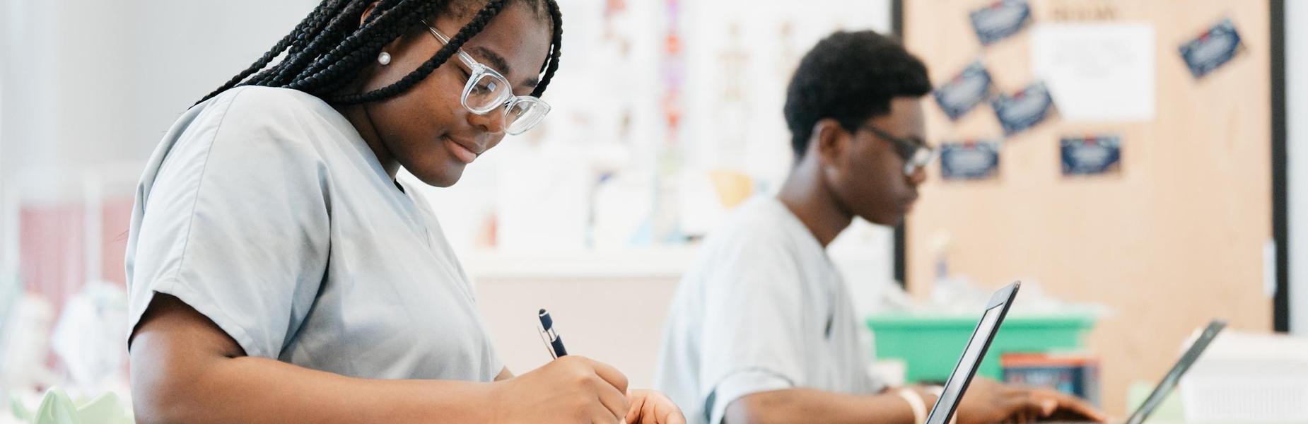 A classroom setting where a student in gray scrubs is writing in a notebook while seated at a green desk. Another student in similar attire is working on a laptop at a nearby desk. The background shows a door with colorful cards attached and a wall with bright educational posters and charts. The room is well-lit with natural light coming from the left side.