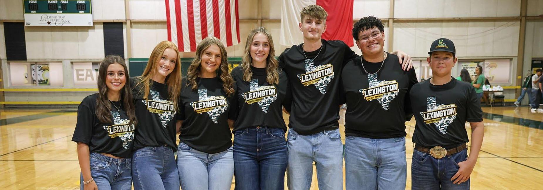 Six people wearing black shirts and wide jeans, standing in a gymnasium.