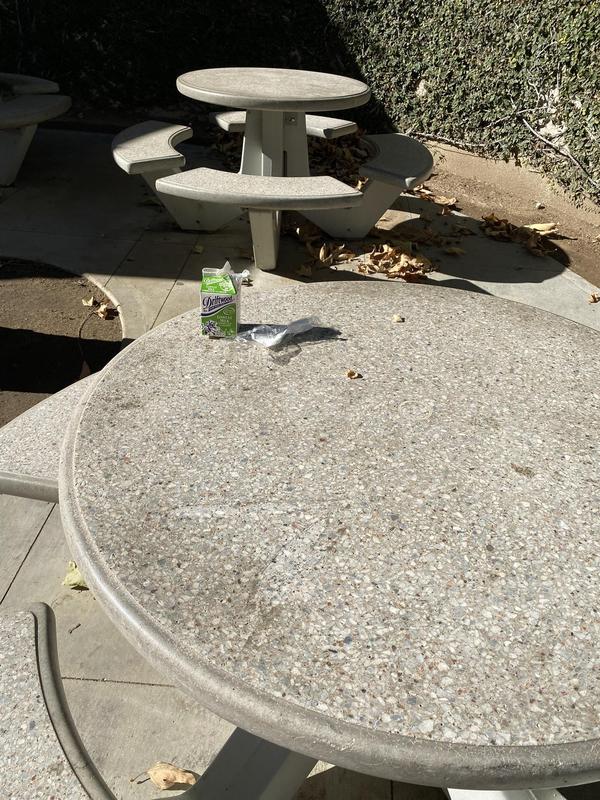 A speckled concrete picnic table top in bright sunlight, with a trash of green milk carton and plastic spoon on its surface
