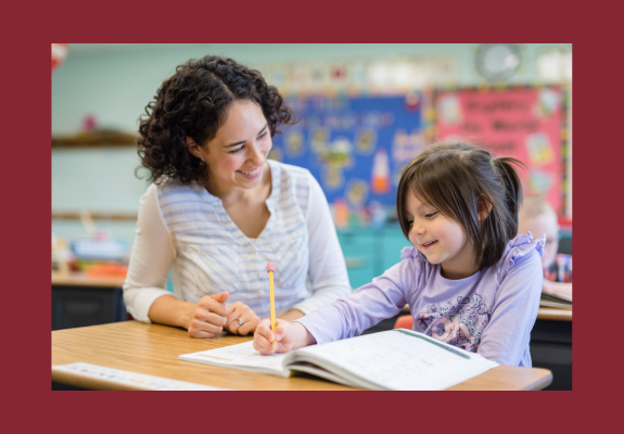 teacher smiles as she helps an elementary student at her desk