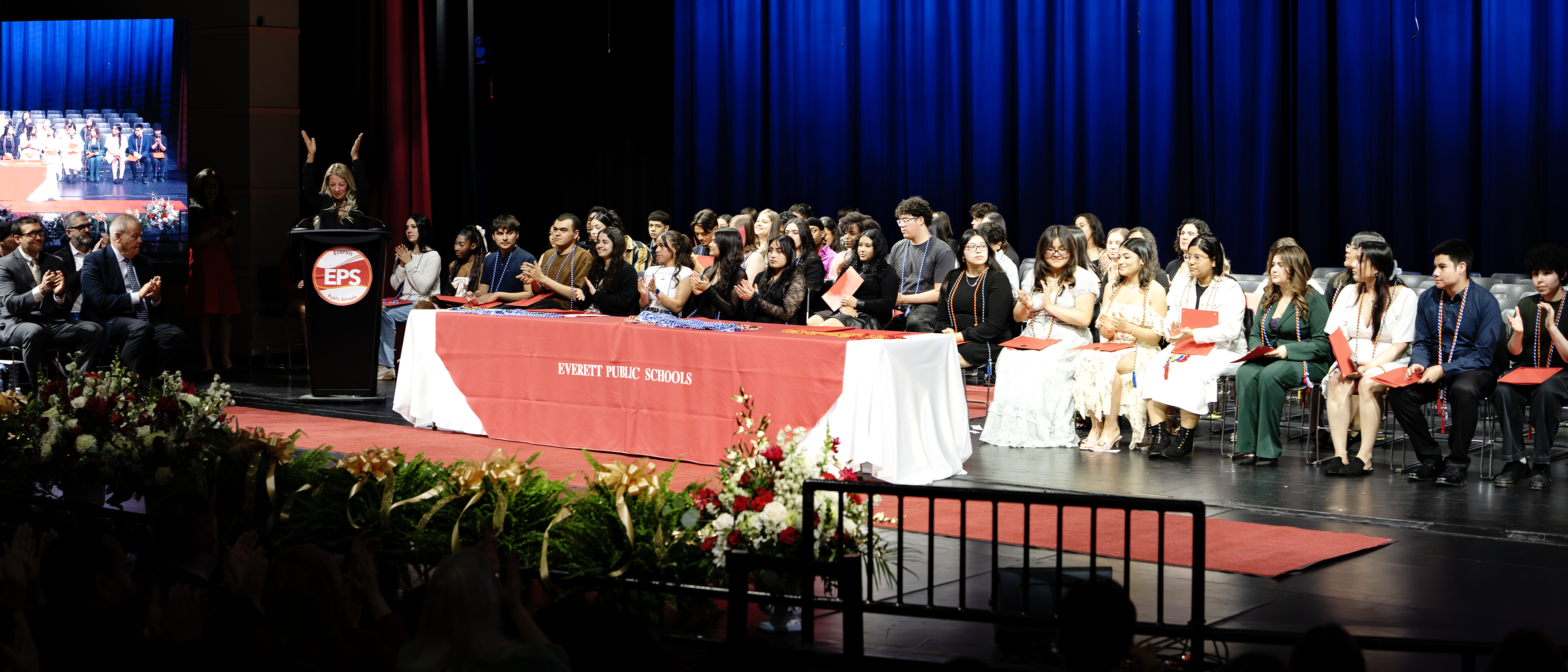 A graduation ceremony with a speaker, audience, and students on stage.
