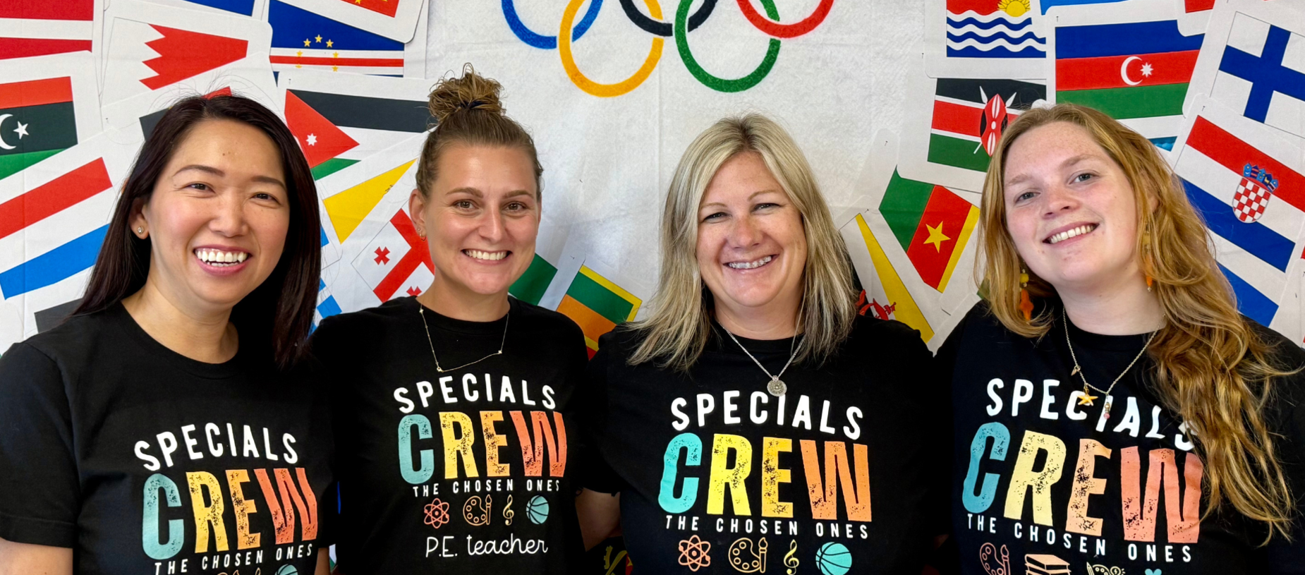 Four women smiling together in themed t-shirts, with a backdrop of flags and Olympic rings.