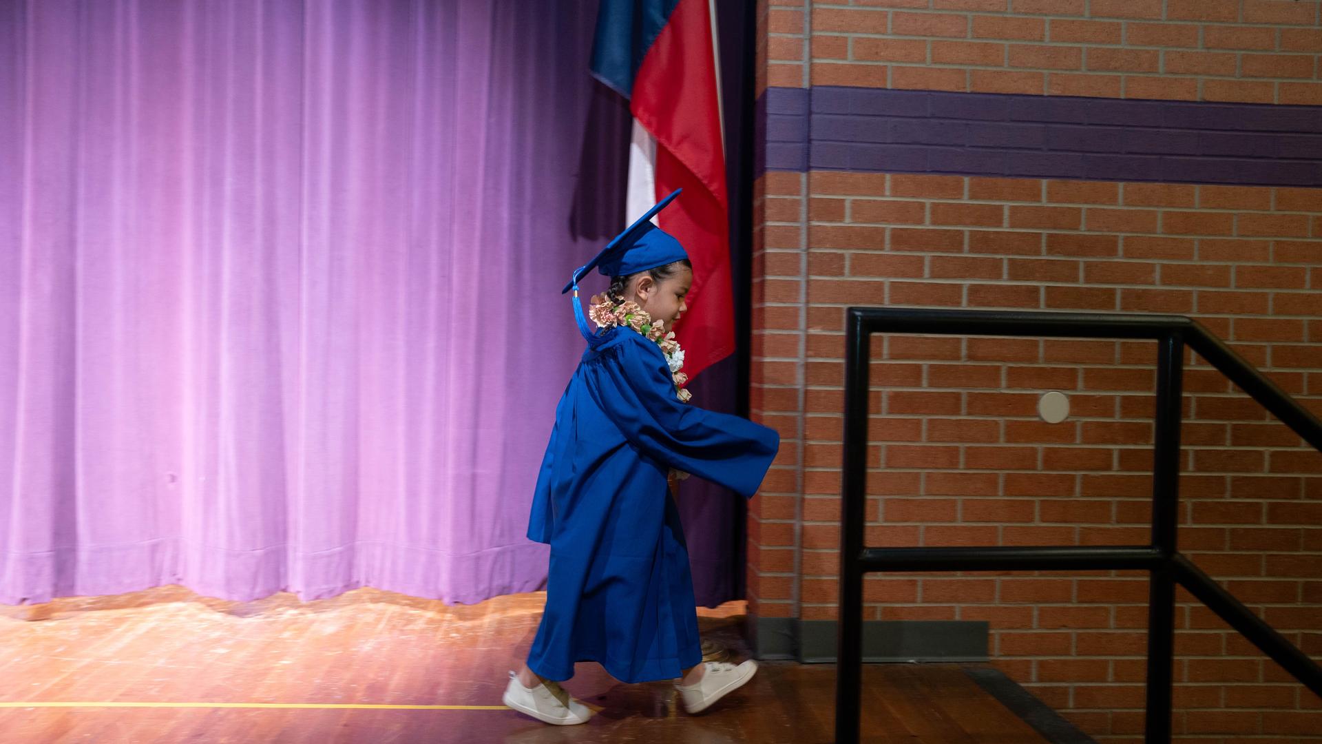 A child in a blue graduation gown walks across a stage with purple curtains.