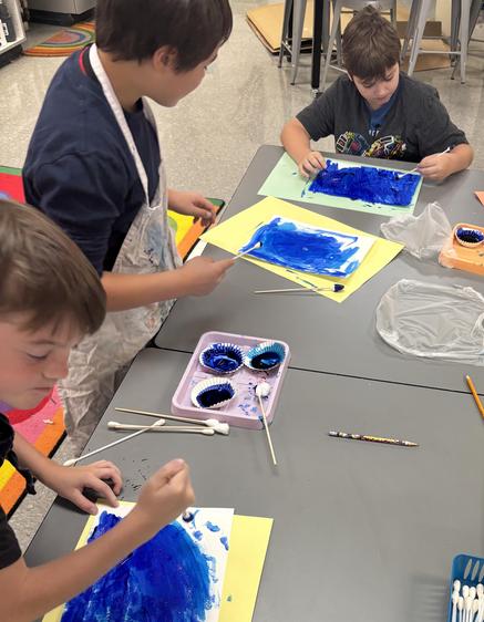 Kids painting on paper with blue paint and cotton swabs at a classroom table.