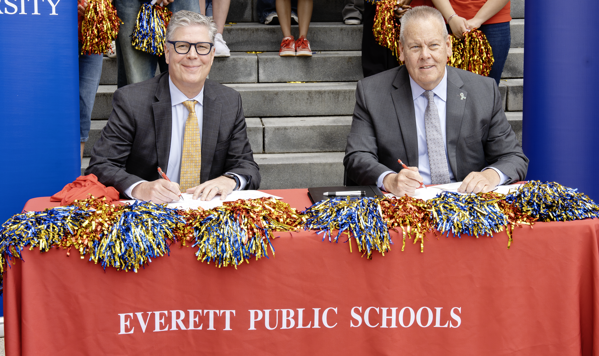Two men sign documents at a table decorated with school colors and pom-poms.