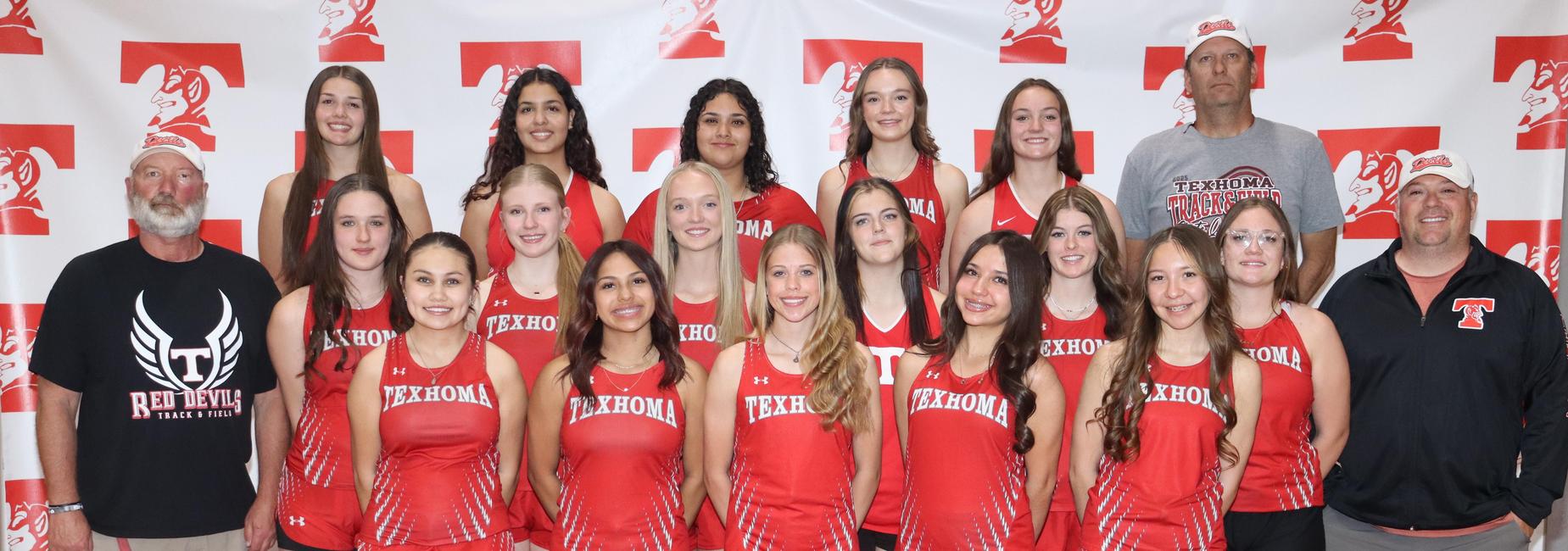 A group of thirteen female athletes in red uniforms stand together in front of a red and white backdrop.