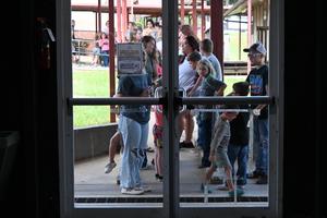 Children and adults waiting in line outside a building, seen through glass doors.