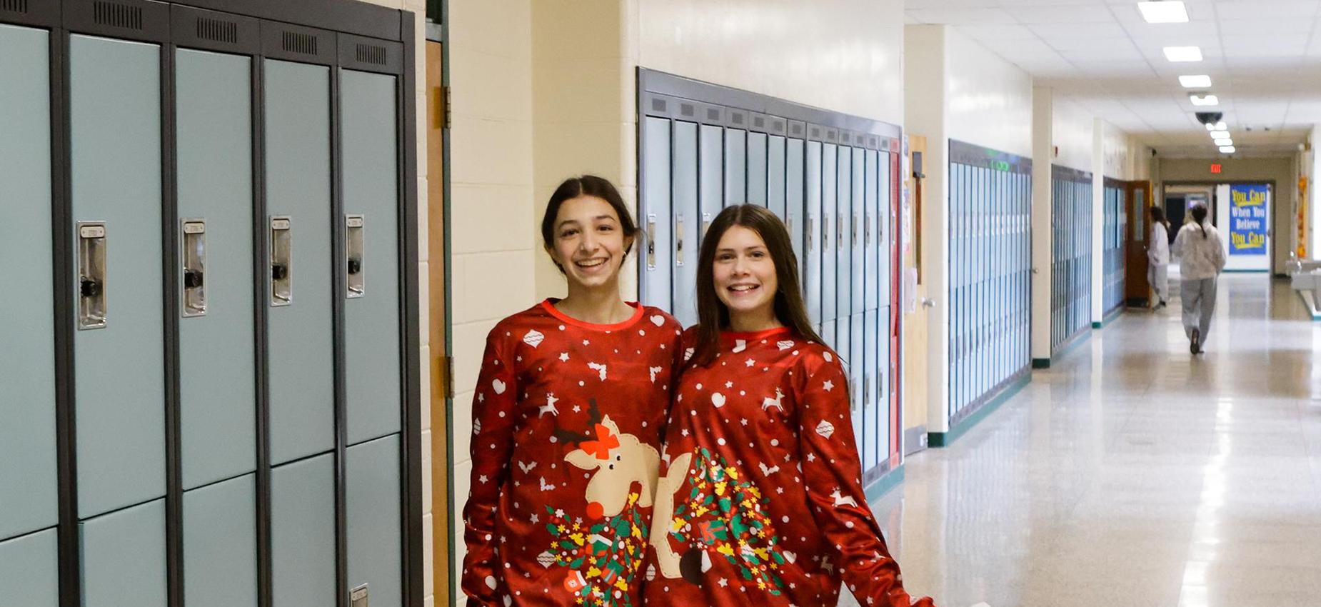 Two girls wearing matching red Christmas sweaters stand in a school hallway.