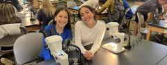 Two girls smile while posing with microscopes at a lab table.