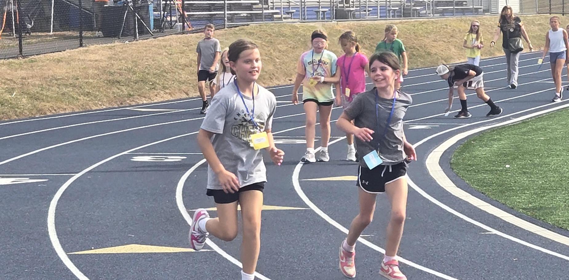 Two girls jogging on a track with other participants in the background.