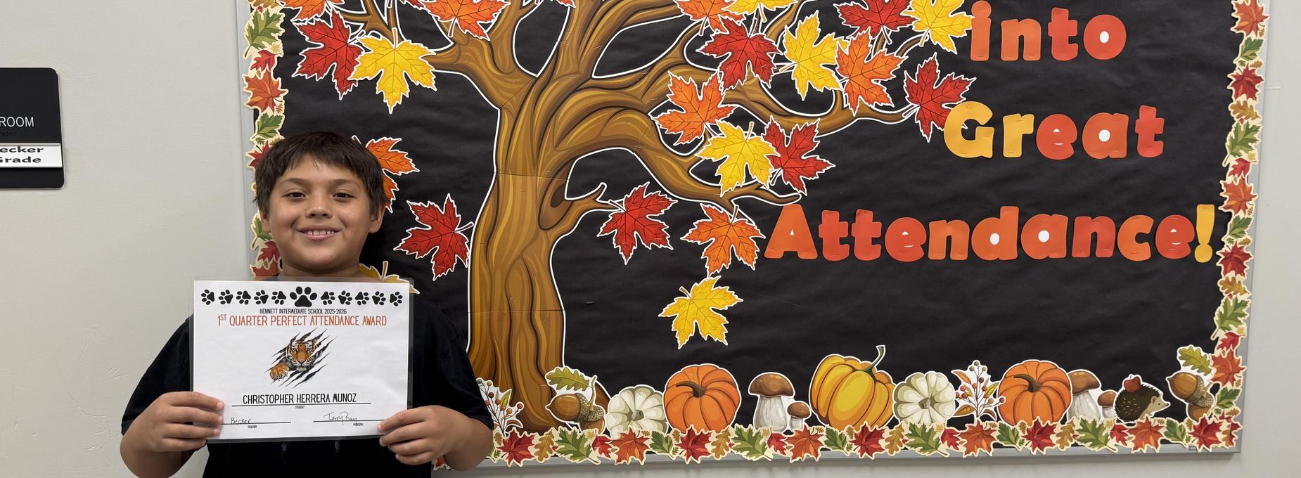 A boy holds a certificate in front of a colorful autumn-themed bulletin board.