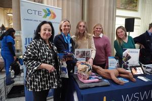 Commission Betty Rosa with instructor and 3 students at BOCES Expo. Table is set with promotional items, including a dog and rabbit.