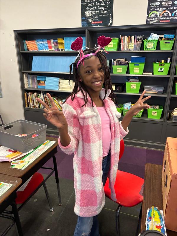 A scholar dressed in pink and read with a hearts headband holding up peace signs