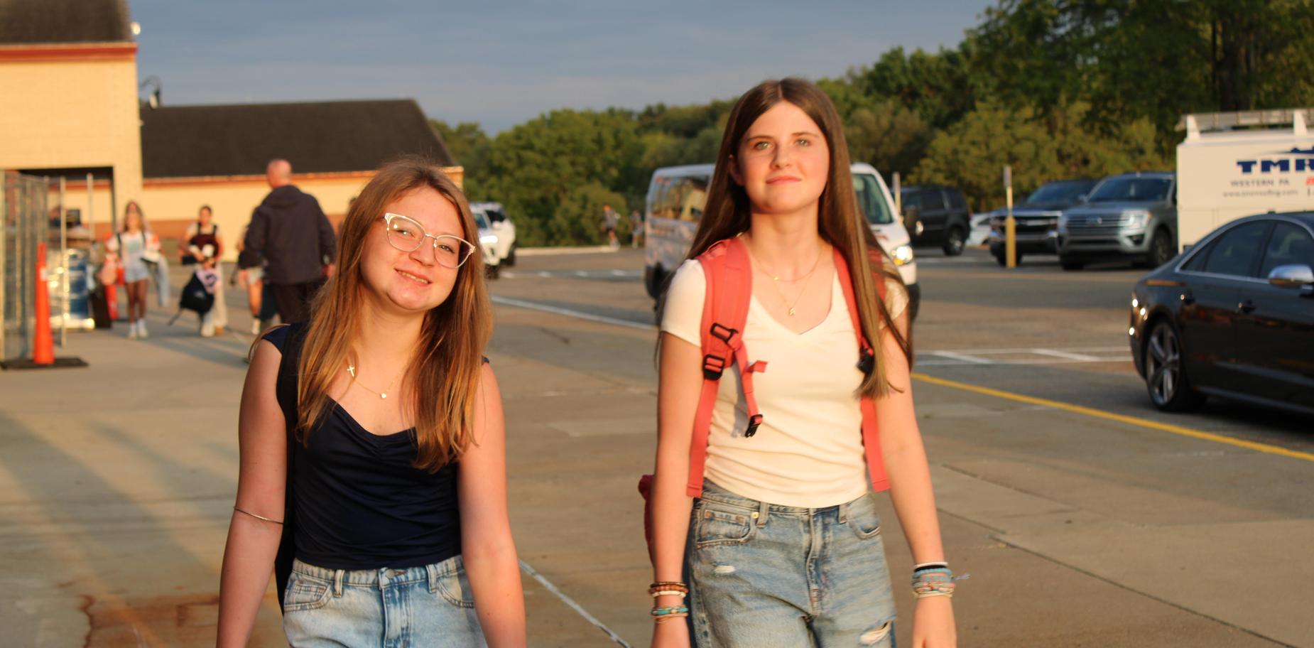Two girls walking together on pavement, wearing casual outfits and smiles.
