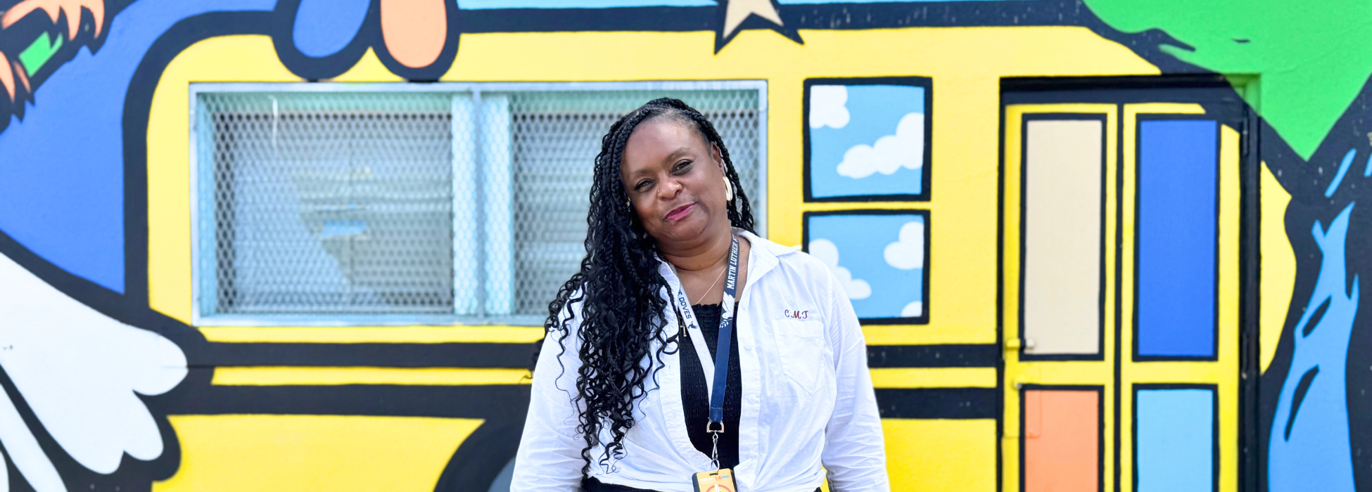 A woman with long curly hair stands in front of a colorful mural featuring a bus.