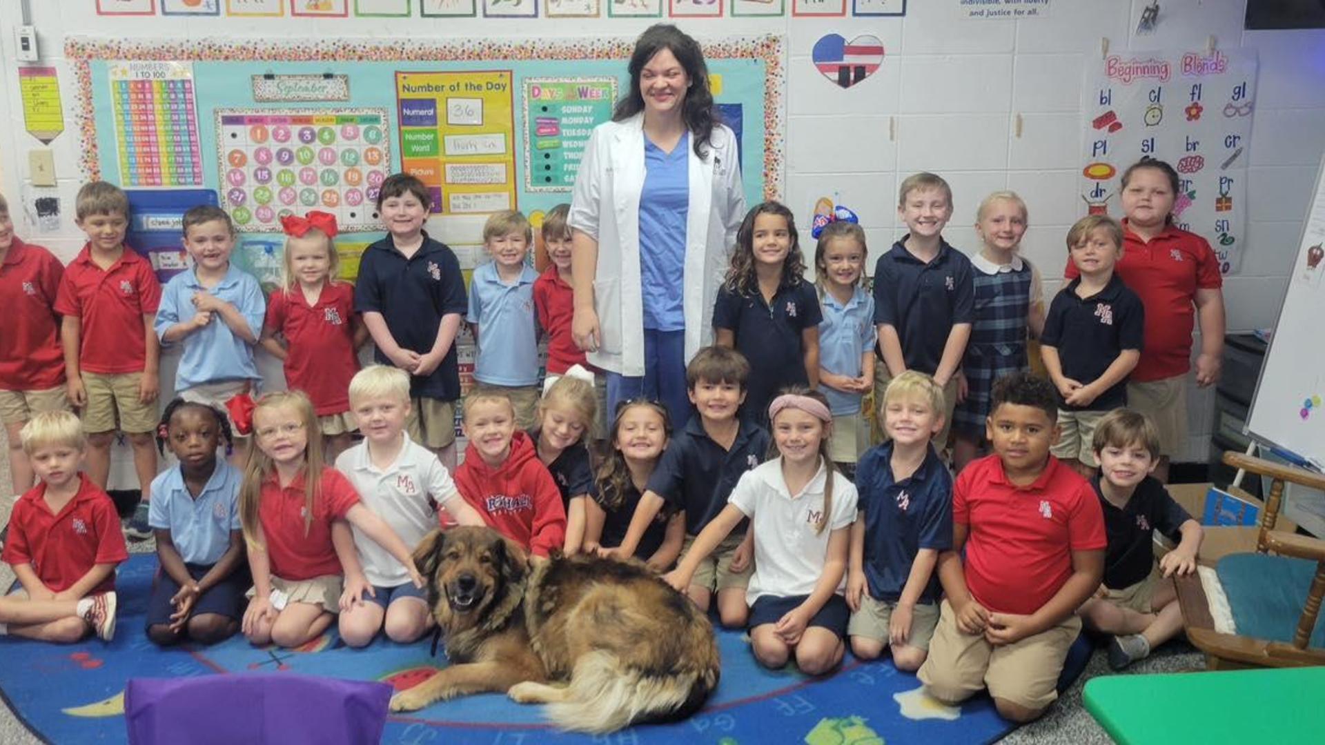 A group of children and a teacher pose together in a classroom with a dog.