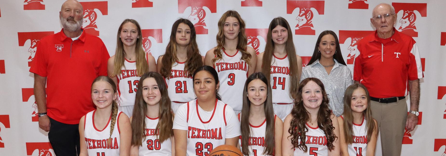 A group of female basketball players posing together with a basketball in front of a team banner.