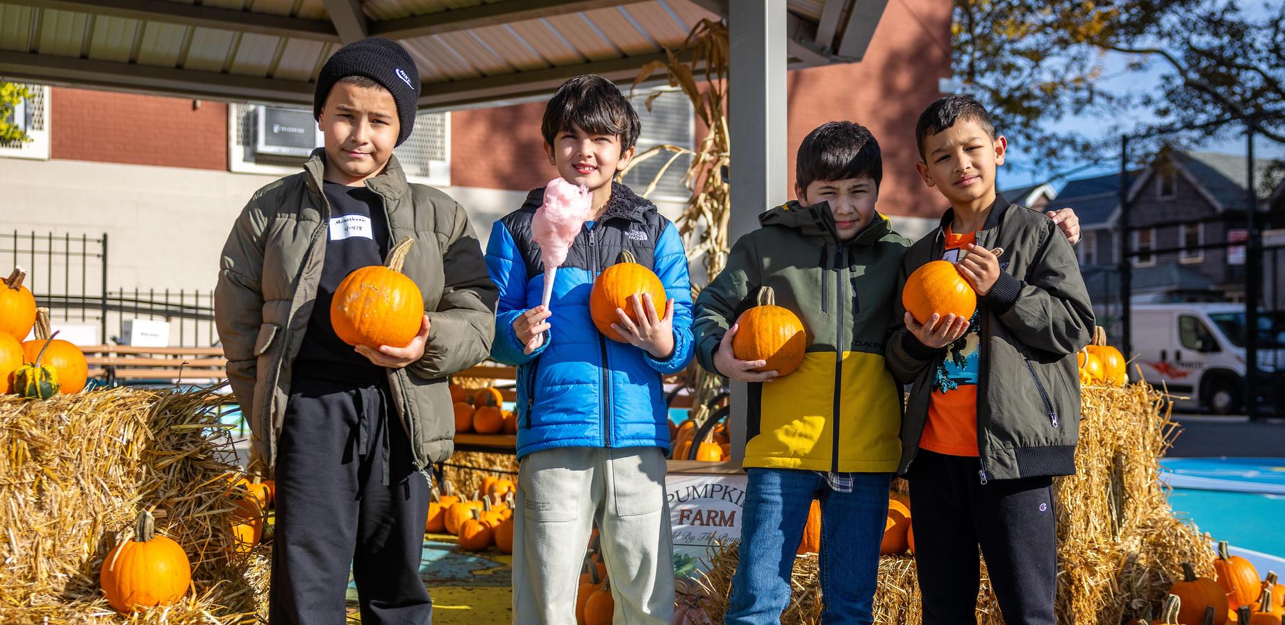 Four children holding pumpkins in a festive farm setting with hay bales.