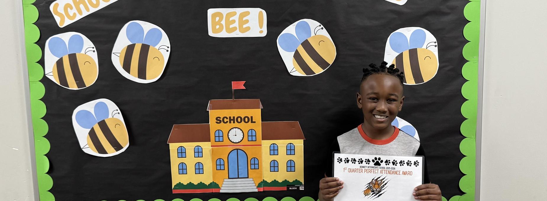 Child smiling beside a bee-themed school decoration, holding a certificate.