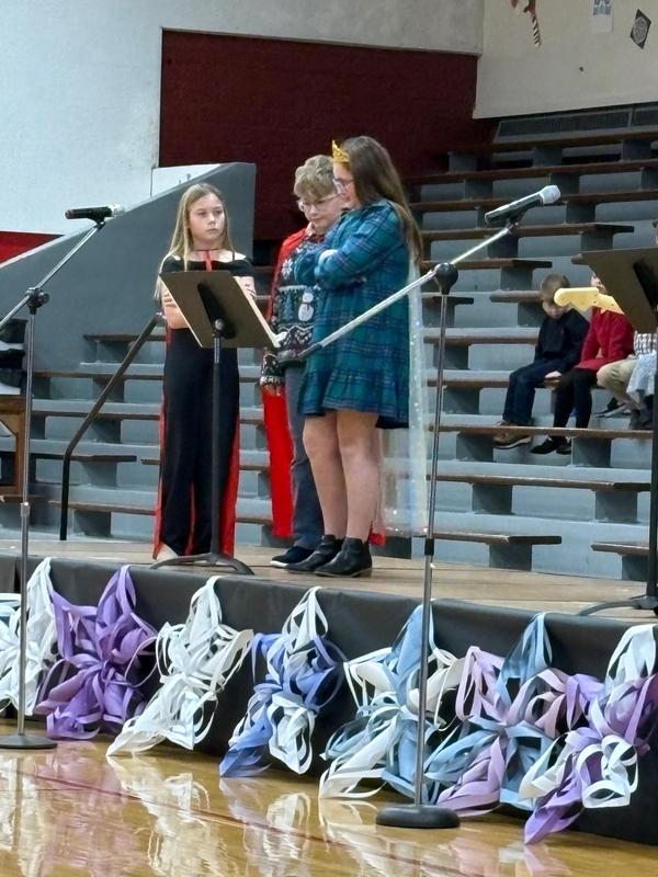 Three children on stage reading from a script during a school event.
