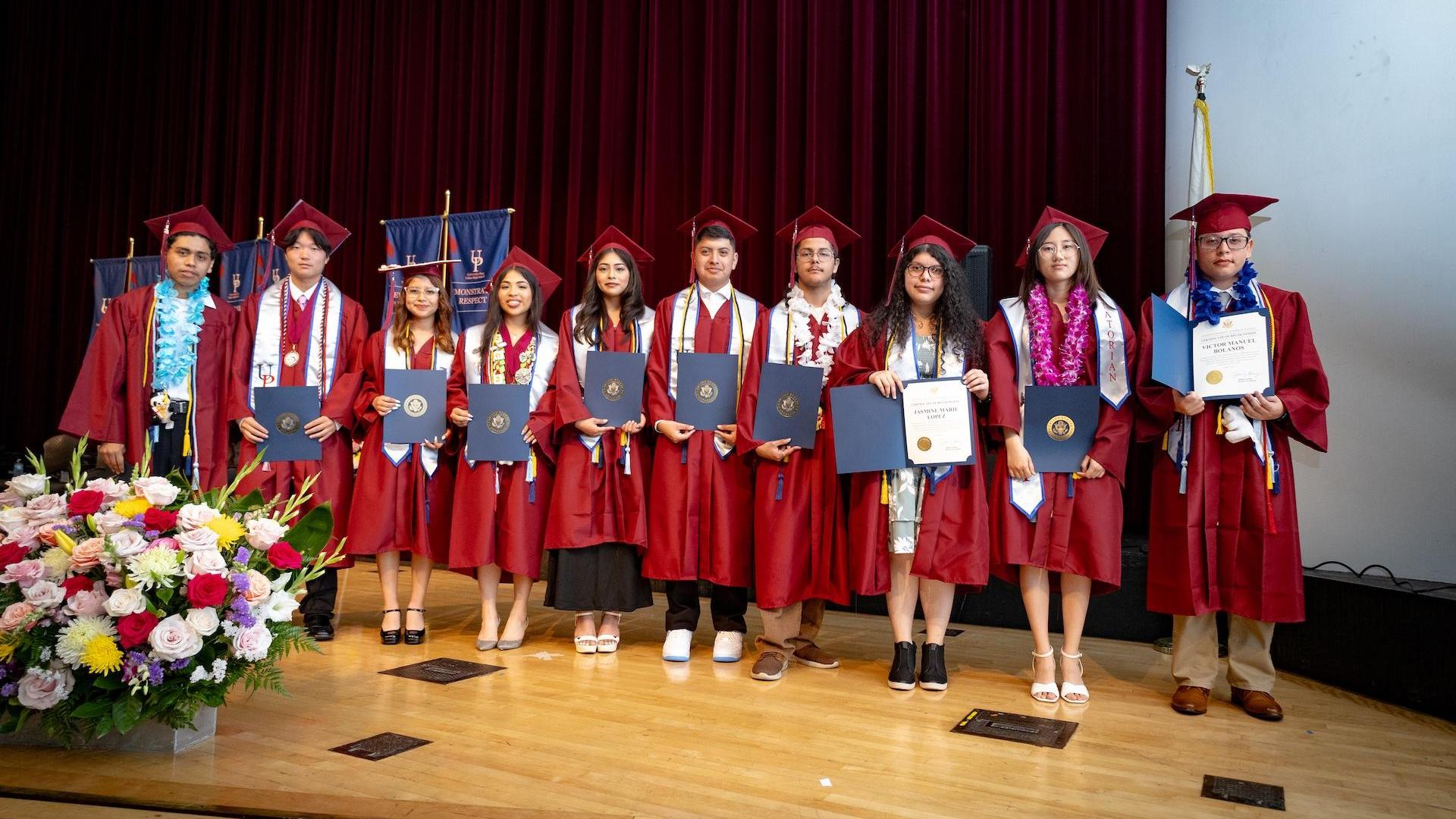 Graduates in gowns holding diplomas on stage with floral arrangement.