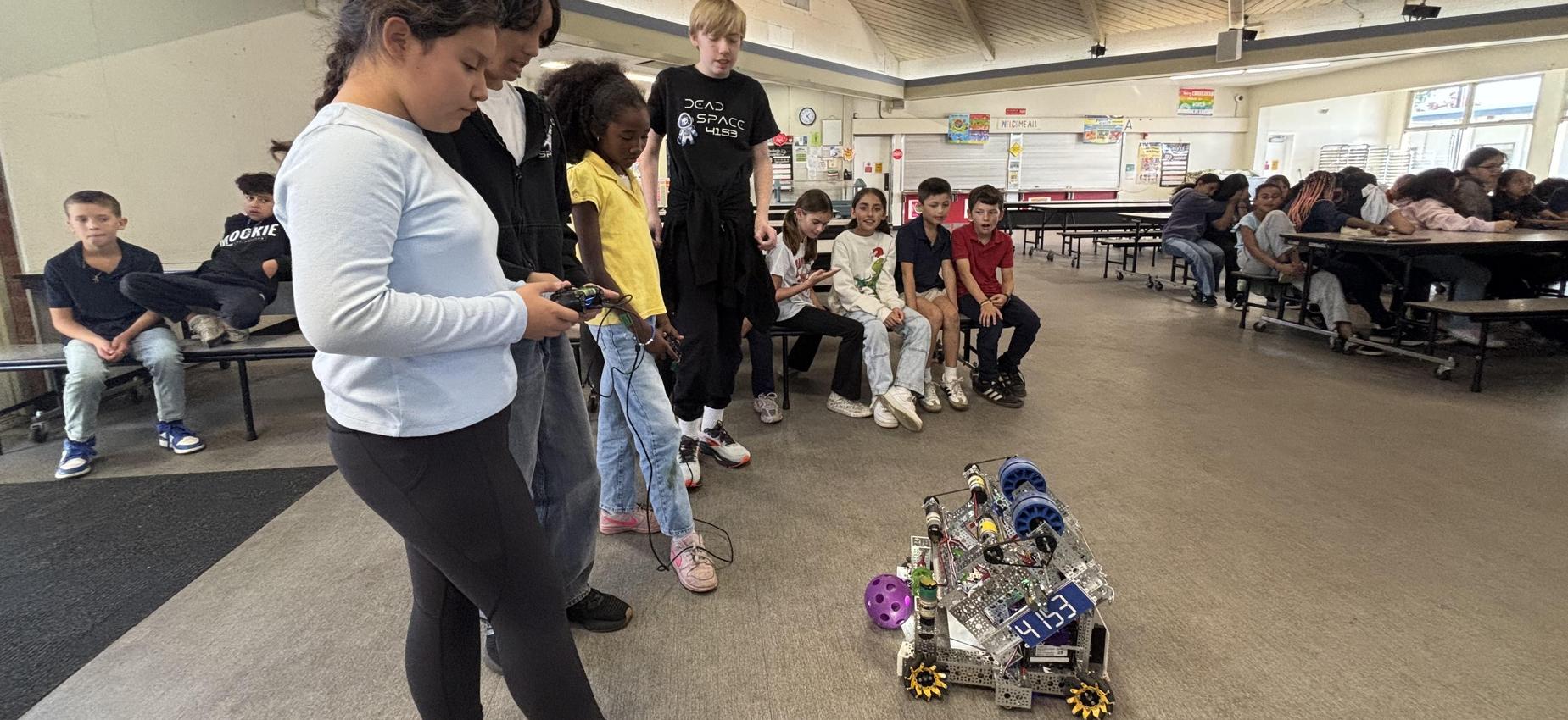 Students controlling a robot during a demonstration in a school cafeteria setting.