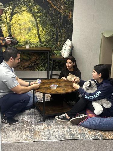 Two people playing cards seated at a table, with a third person taking a photo.