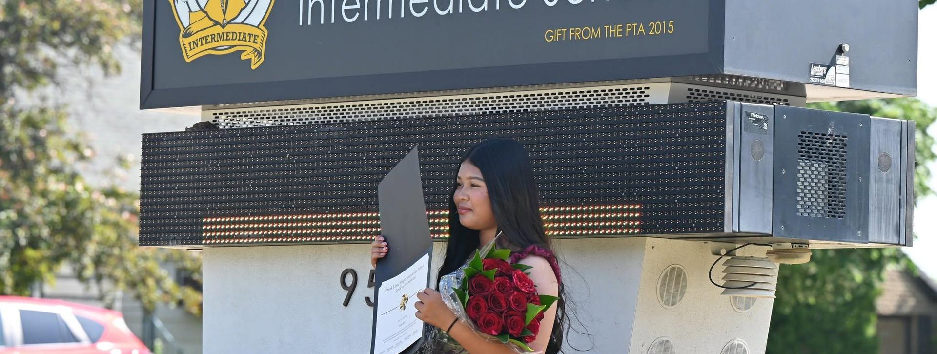 A young woman holding a bouquet of flowers and a certificate stands by a school sign.
