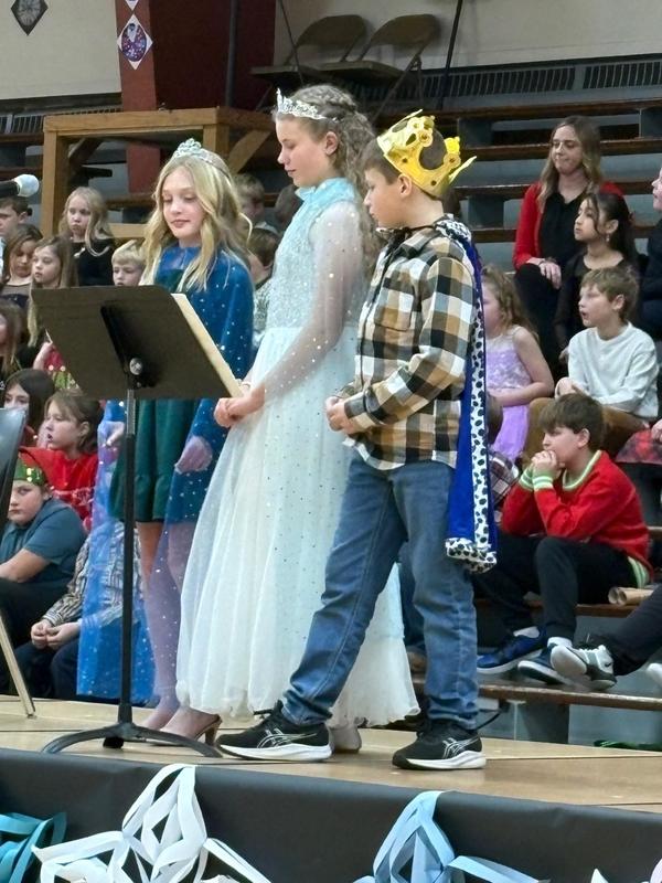 Three children in costumes standing on stage while a crowd watches in the background.