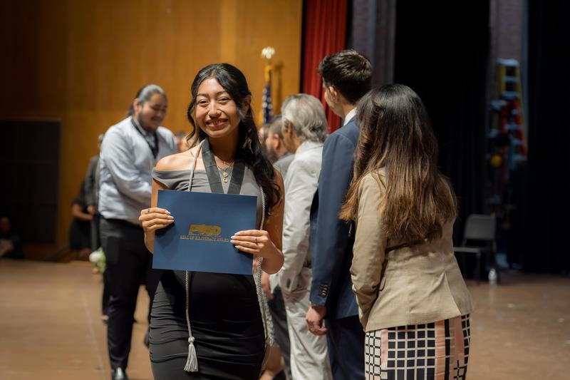 A student in a black dress smiles while holding a PUSD Seal of Biliteracy Award folder on stage, wearing a silver cord and medal as others line up behind her.
