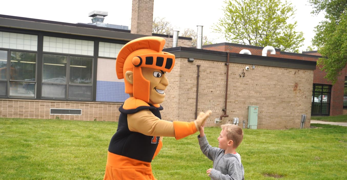 The Trojan mascot greets a smiling boy outside a school building.